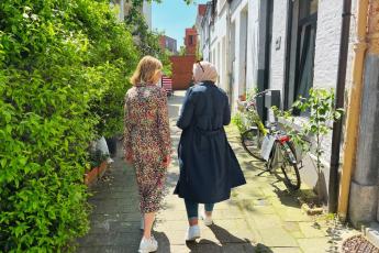 Grete en Fatima, 2 vrouwen, wandelen door een straat. Ze wandelen met hun rug naar de camera. De zon schijnt. Grete is een blonde vrouw en draagt een kleurige jurk. Fatima draagt een witte hoofddoek en een blauwe jas.